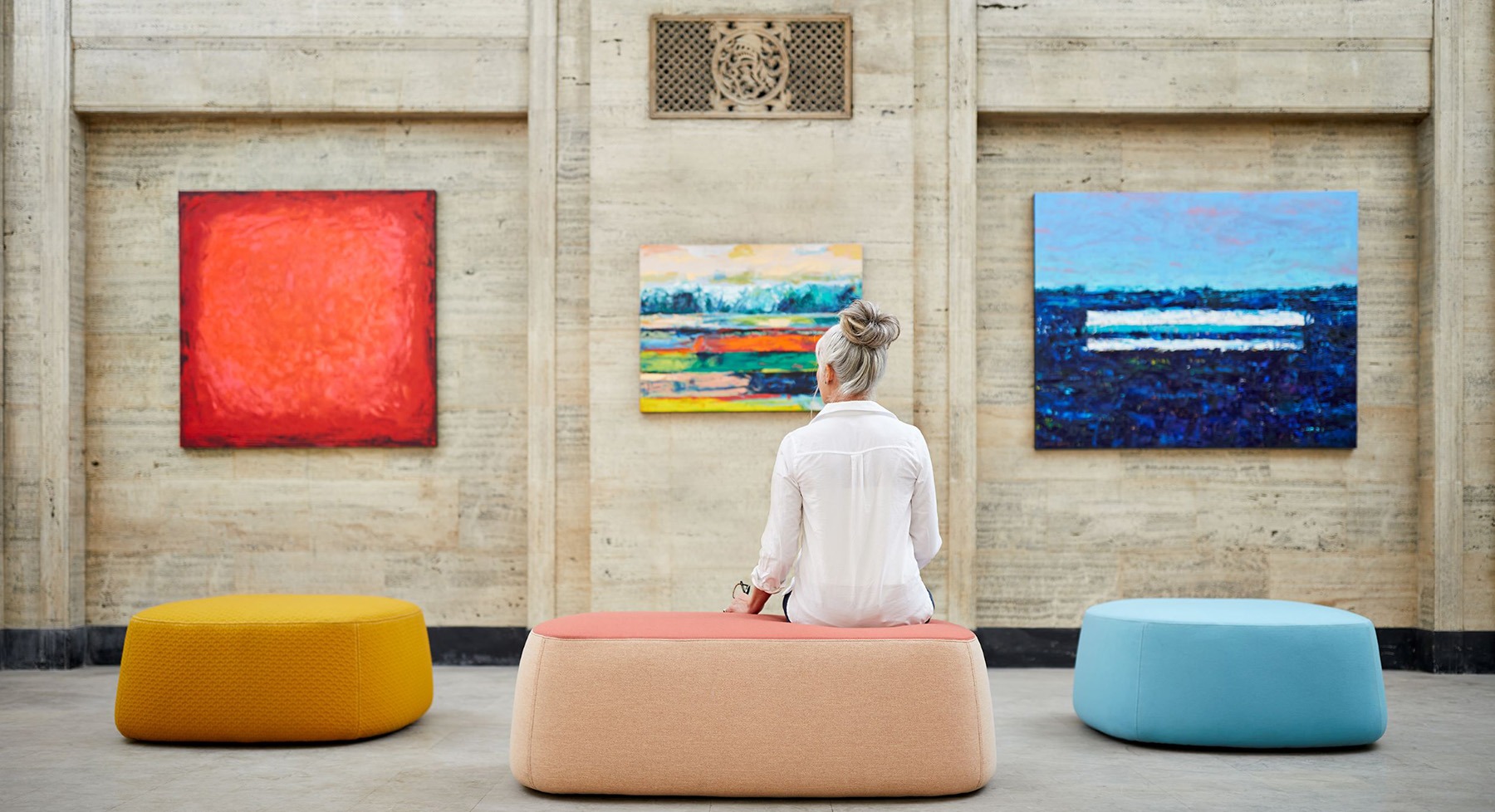 une femme assise sur un pouf regardant trois tableaux au mur